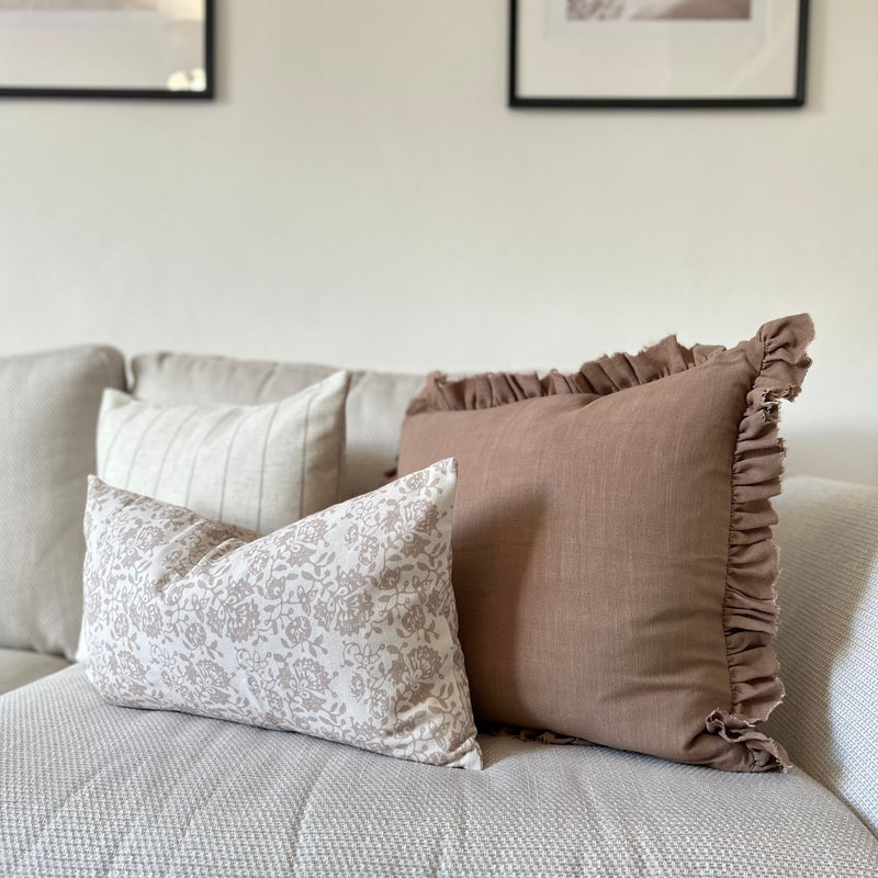 set of 3 cushions. 1 Square brown with ruffled edge, 1 Square Cream cushion with a thin vertical brown stripe and one rectangle cream cushion with a brown floral pattern sat in front of the 2 other cushions. All sat on a cream sofa