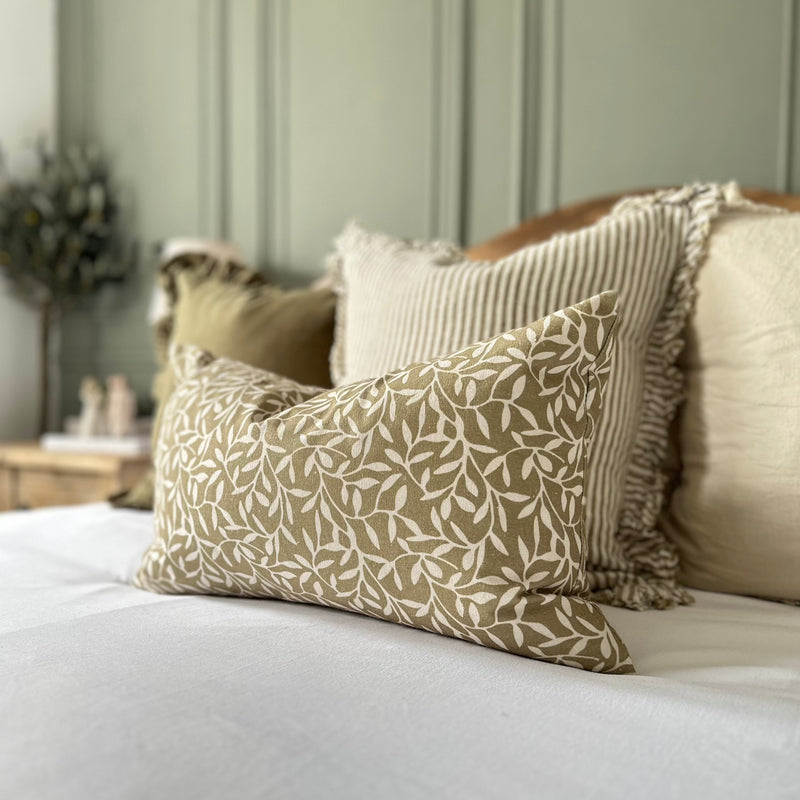 Clos up olive rectangle cushion with cream olive leaves print. Sat on white bedding . With green tones around the room.