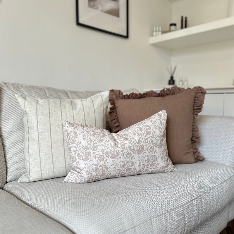 set of 3 cushions. 1 Square brown with ruffled edge, 1 Square Cream cushion with a thin vertical brown stripe and one rectangle cream cushion with a brown floral pattern sat in front of the 2 other cushions. All sat on a cream sofa