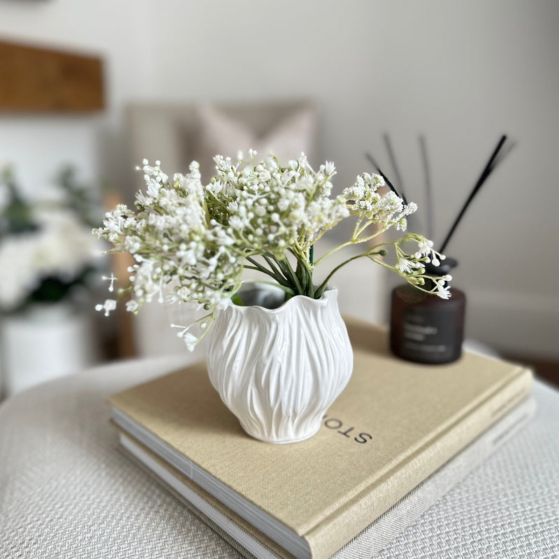 white ceramic vase in a tubular floral shape with a ribbed effect edge. filled with white baby breathe flowers stood on a stack of books