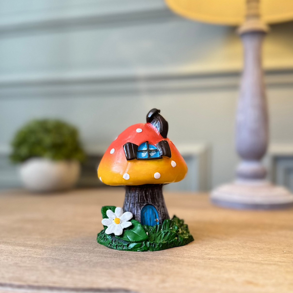 Ceramic toadstool incense burner. orange top with white polka dots. Base is like a tree trunk with a little blue door. Sat on a grassy base with a white daisy.