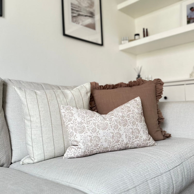 set of 3 cushions. 1 Square brown with ruffled edge, 1 Square Cream cushion with a thin vertical brown stripe and one rectangle cream cushion with a brown floral pattern sat in front of the 2 other cushions. All sat on a cream sofa