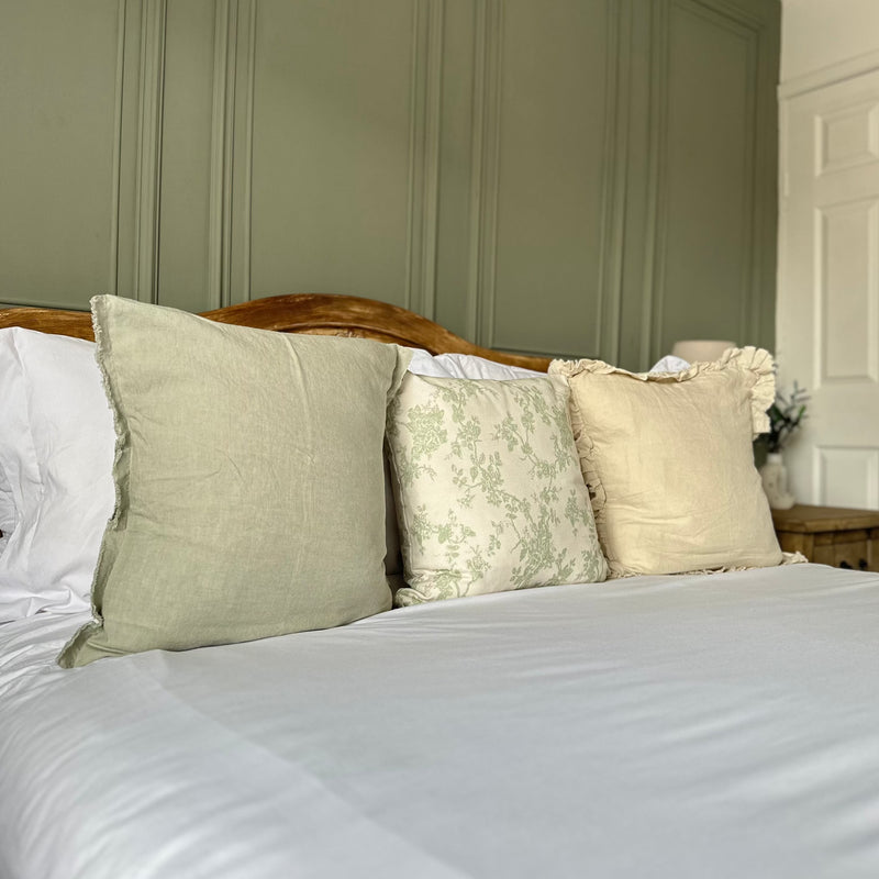 set of 3 square cushions. 1 sage green, 1 cream with delicate green floralprint and 1 cream with a ruffled edge. Sat on a bed with white bedding and a green back wall.