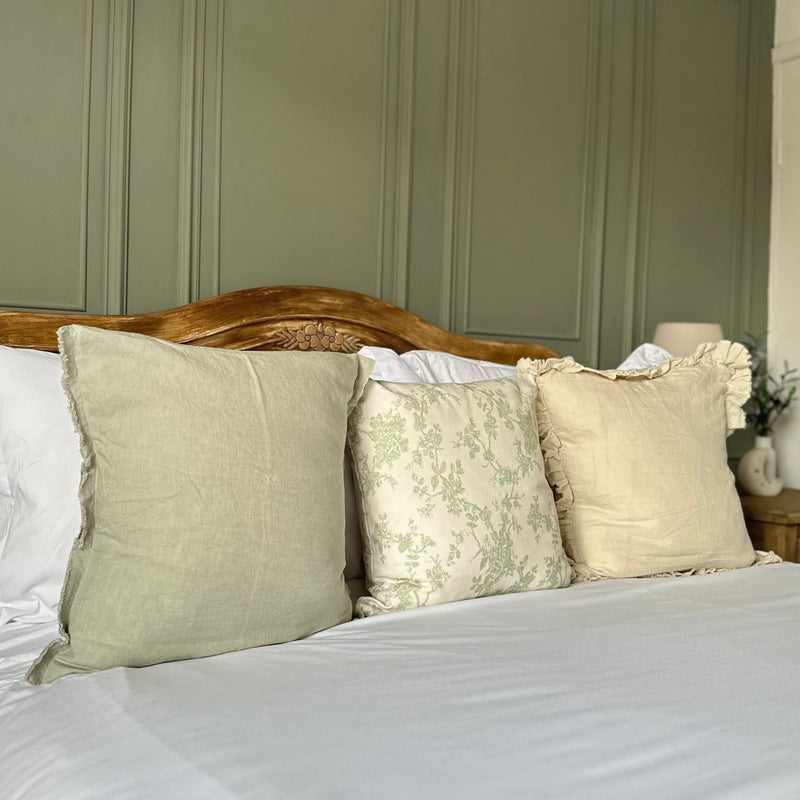 set of 3 square cushions. 1 sage green, 1 cream with delicate green floralprint and 1 cream with a ruffled edge. Sat on a bed with white bedding and a green back wall.