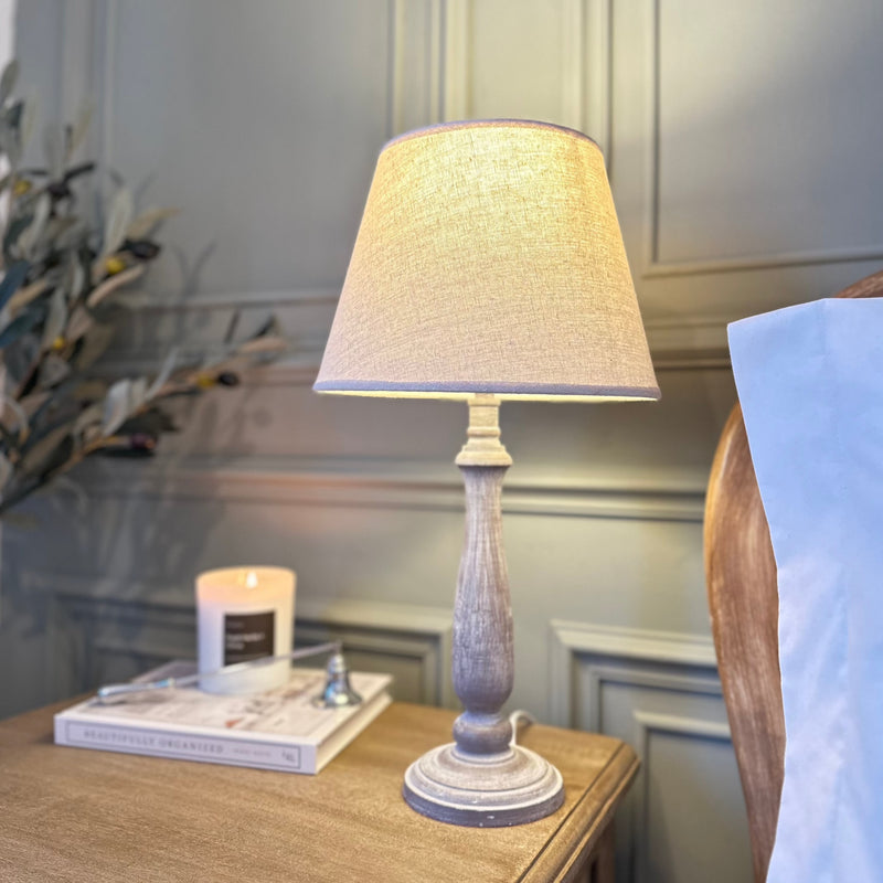 slim wooden white wash table lamp with a cream linen shade, sat on a wooden bedside table with a dark green panelled wall as a backdrop in a country style setting.