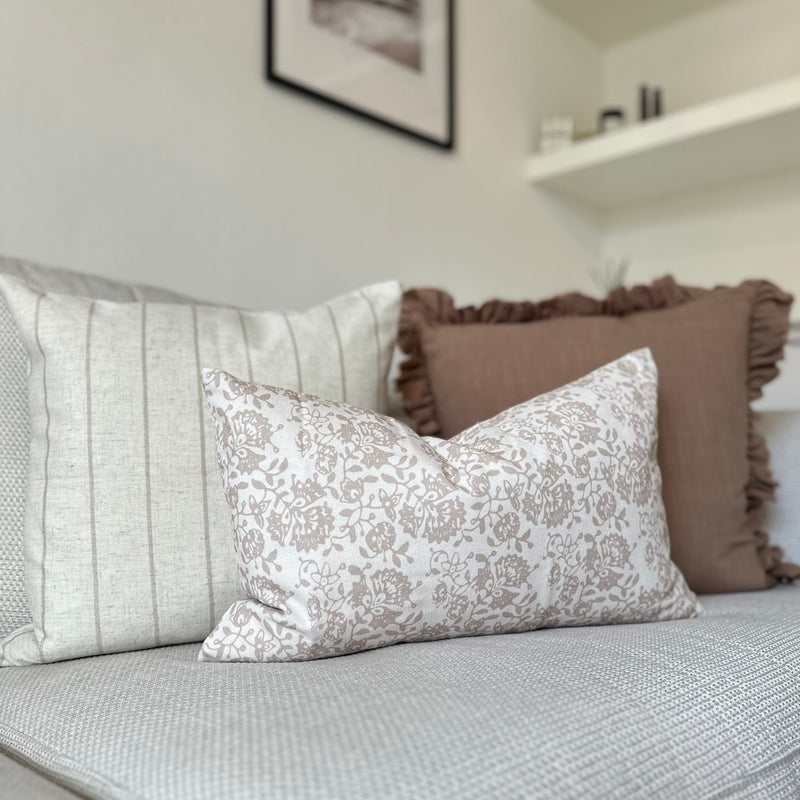 set of 3 cushions. 1 Square brown with ruffled edge, 1 Square Cream cushion with a thin vertical brown stripe and one rectangle cream cushion with a brown floral pattern sat in front of the 2 other cushions. All sat on a cream sofa