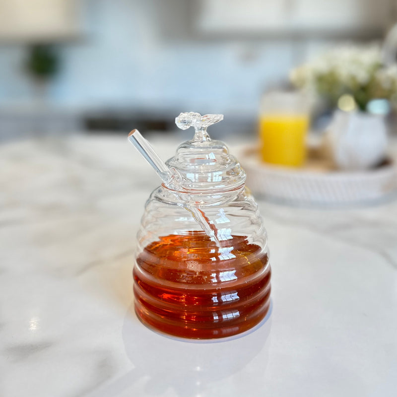 glass honey jar with glass dropper and glass lid with glass bumble bee on top. Jar full of honey. All sat on a white kitchen counter