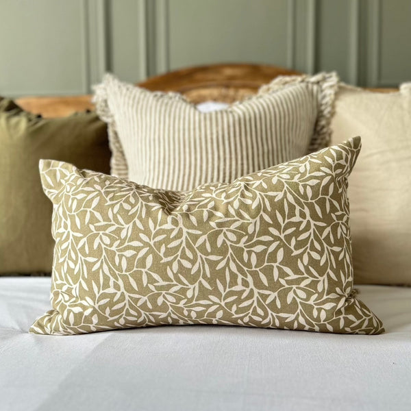 olive coloured rectangle cushion wit a cream olive leaf print. Sat on a bed with white bedding and panelled olive green walls as a back drop.