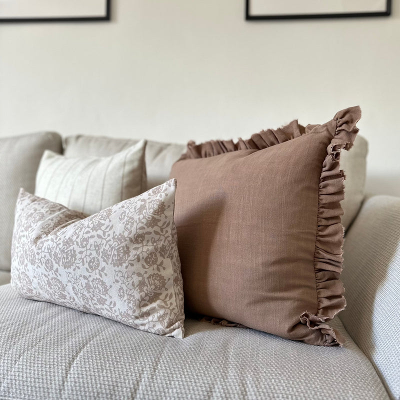 Side angle of a Set of 3 cushions. 1 Square brown with ruffled edge, 1 Square Cream cushion with a thin vertical brown stripe and one rectangle cream cushion with a brown floral pattern sat in front of the 2 other cushions. All sat on a cream sofa