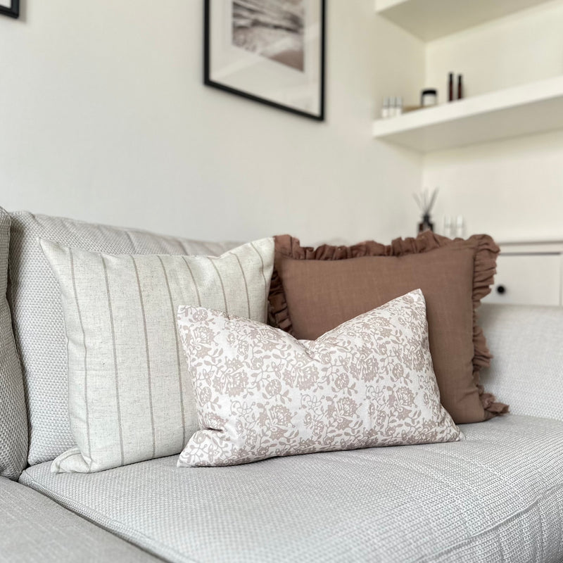 set of 3 cushions. 1 Square brown with ruffled edge, 1 Square Cream cushion with a thin vertical brown stripe and one rectangle cream cushion with a brown floral pattern sat in front of the 2 other cushions. All sat on a cream sofa