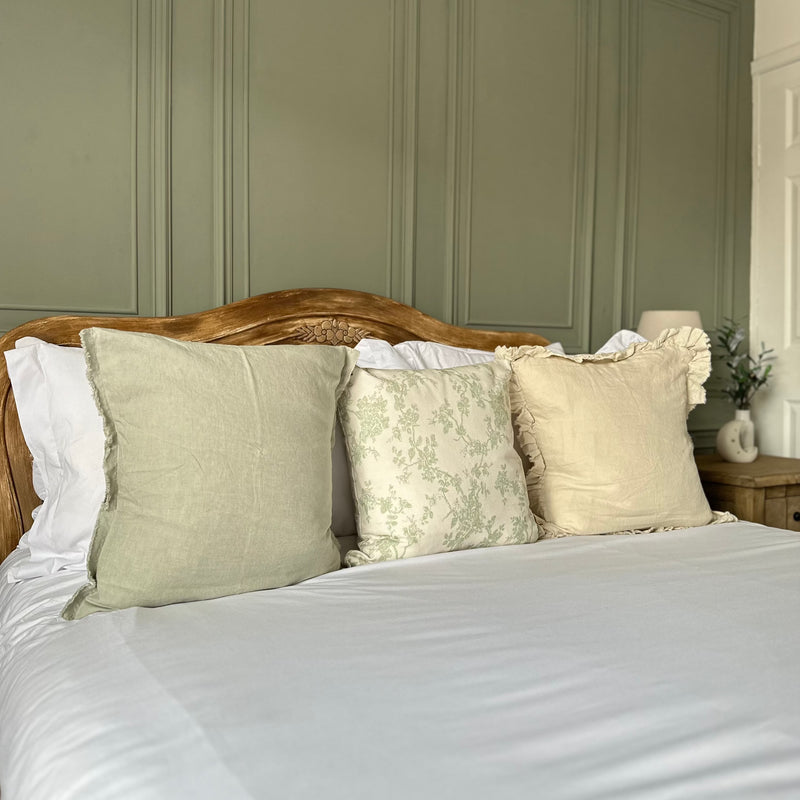 set of 3 square cushions. 1 sage green, 1 cream with delicate green floralprint and 1 cream with a ruffled edge. Sat on a bed with white bedding and a green back wall.