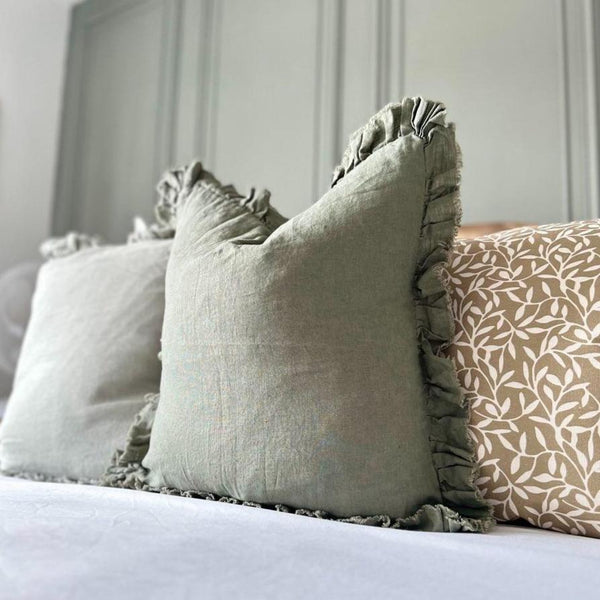 Ruffled Edge Khaki Green Cushion sat on a bed with a green back wall 