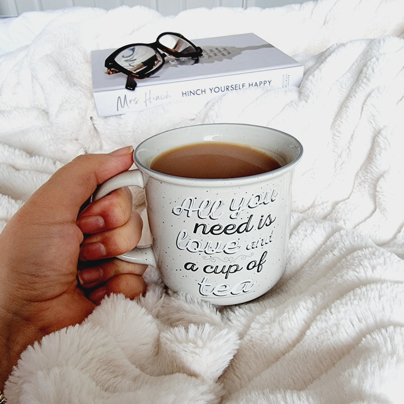 Lady holding mug on a fluffy white throw. Mug is light grey with "All you need is love and a cup of tea" written on front in white and dark grey writing
