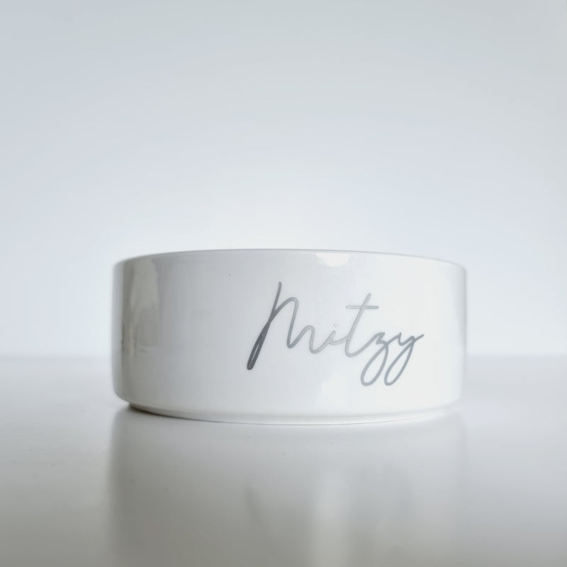 White ceramic pet bowls on white background with "Mitzy" written on bowl in silver script font