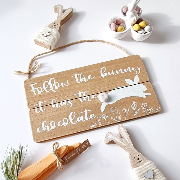 brown wooden plaque with "Follow the bunny it has the chocolate! written in white with a jumping bunny printed in white with a white 3D pom pom tail. All on a white background surrounded by Easter ornaments 