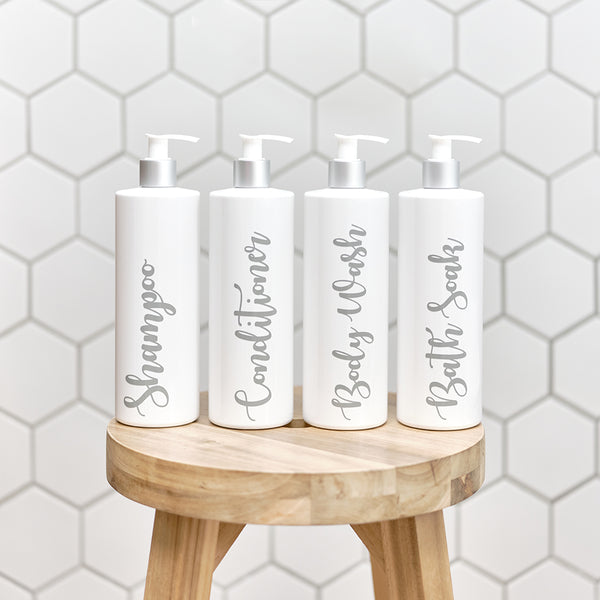 Four sleek and stylish 500ml white and silver reusable dispenser pump bottles with customised grey wording for Shampoo, Conditioner, Body Wash, and Bath Soak. Bottles are placed on a wooden stool in a bathroom setting.