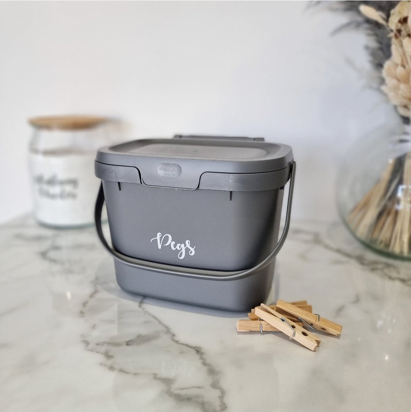 Dark grey storage caddy with lid and carry handle. With pegs written on front in white, sat on white and grey marble counter top