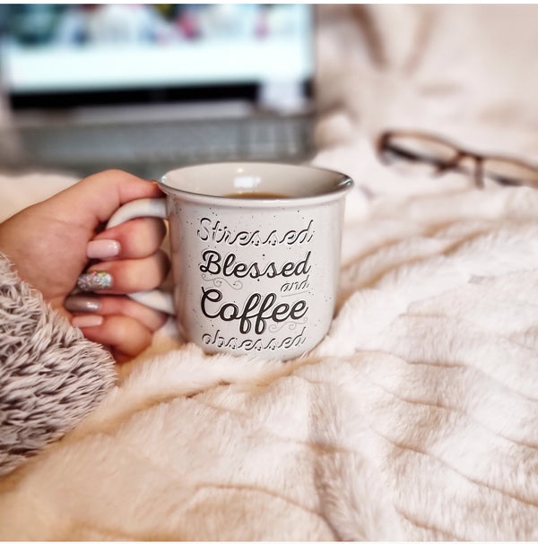 lady holding light grey ceramic mug on a fluffy white throw, mug has "Stressed Blessed and Coffee obsessed" written on in white and grey front 